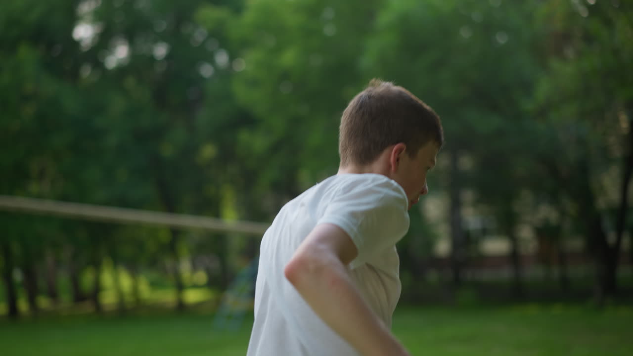 A young boy in a white shirt jumps while trying to maintain balance, with sunlight reflecting from above, the blurred background shows sports equipment and trees