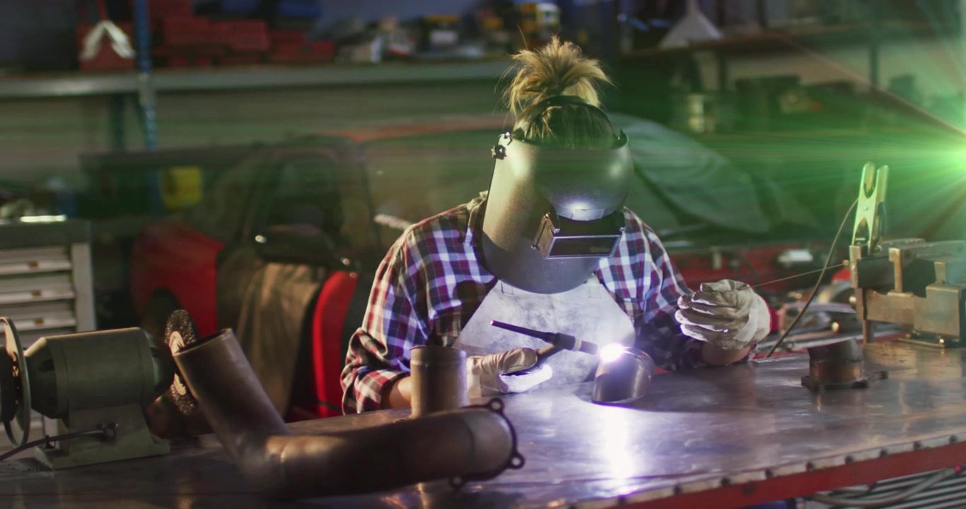 Female welder picking up helmet and welding steel exhaust pipe with sparks in automotive workshop