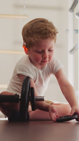 focused little boy puts bumper plates on heavy barbell sitting on wooden floor. blond toddler prepares weight equipment for workout in gym slow motion