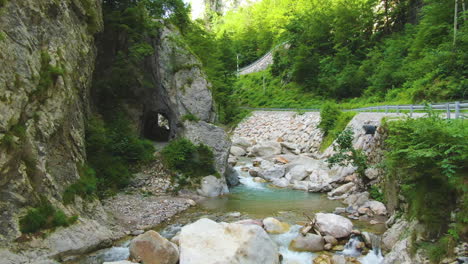 Klarer-Flacher-Fluss-Mit-Steinen,-Umgeben-Von-Felsen,-Grün-Und-Verkehrsstraße