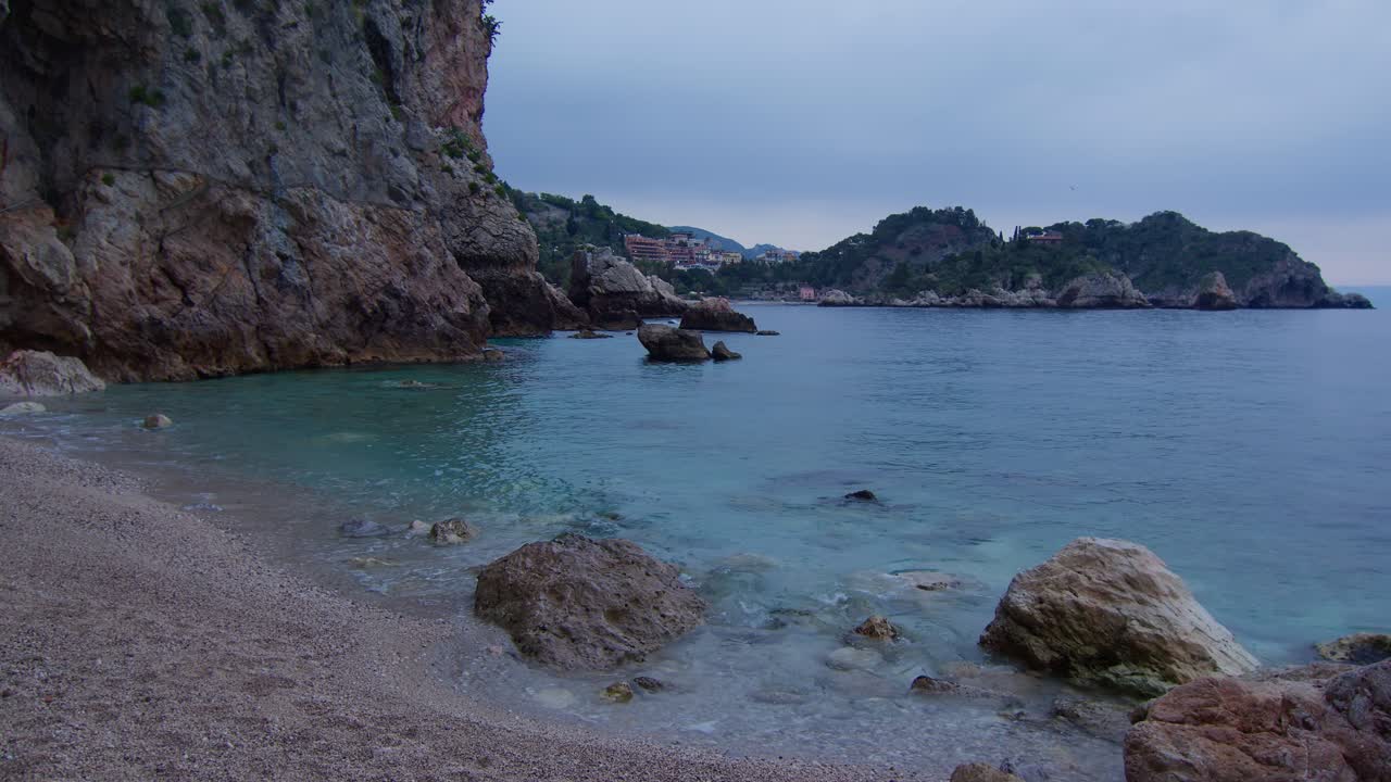Calm sea gently caresses rocky cave beach with Isola Bella on horizon in Taormina, Sicily, Italy (Sicilia, Italia), with quiet White Lotus cinematic mood