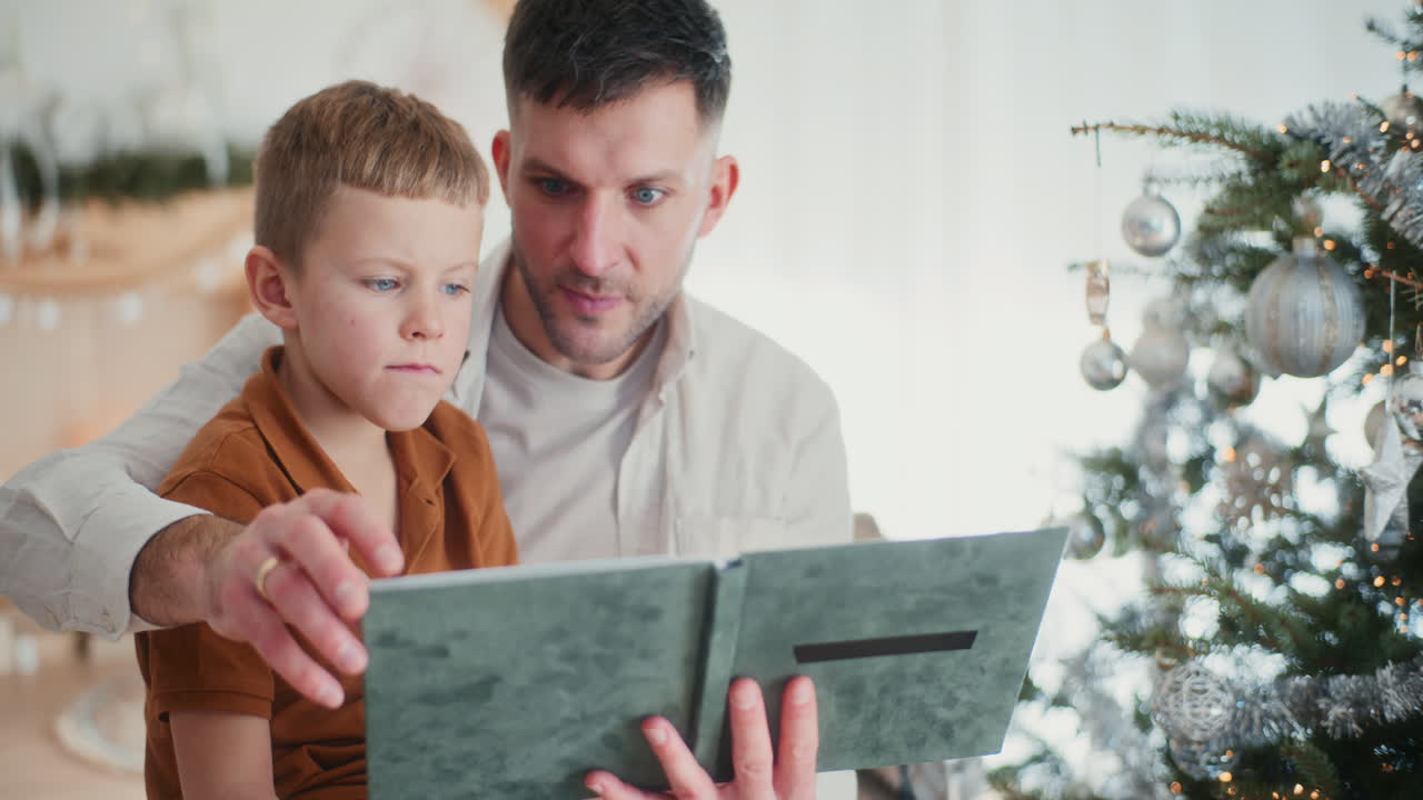 papá y el niño mirando a través de fotos en un álbum durante las vacaciones con decoraciones de navidad en el fondo