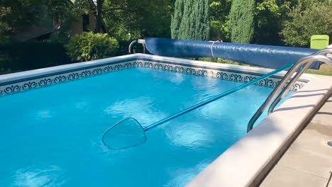 Vista-Panorámica-De-La-Limpieza-De-La-Superficie-De-La-Piscina-Al-Aire-Libre-En-Casa-Con-Un-Skimmer-De-Hojas-De-Malla-Fina-En-Cámara-Lenta-Y-Un-Pintoresco-Jardín-De-Pinos-En-El-Fondo