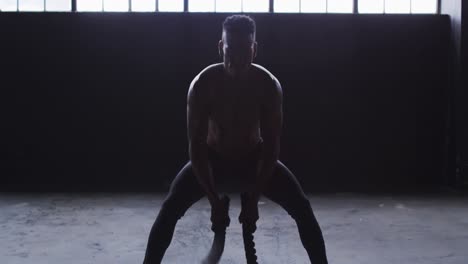 shirtless african american man exercising battling ropes in an empty building