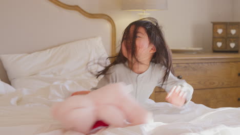 young girl playing on bed wearing pyjamas at home cuddling soft toy