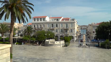 Statische-Ansicht-Des-Hauptplatzes-Von-Syros-In-Richtung-Meer-Mit-Palme,-Griechischer-Flagge-Und-Autos-In-Zeitlupe