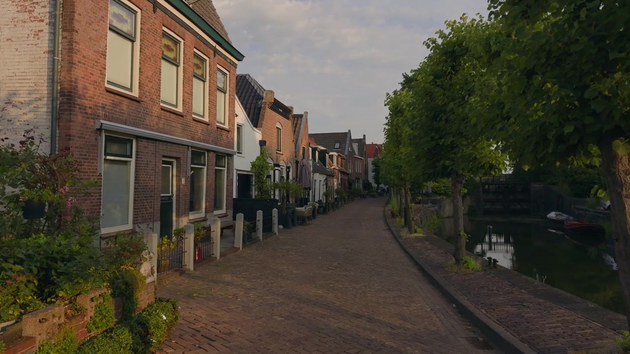 POV (Point of View) gåtur gennem en charmerende hollandsk gade langs en kanal omkranset af træer og historiske huse under en blød blå sommerhimmel.