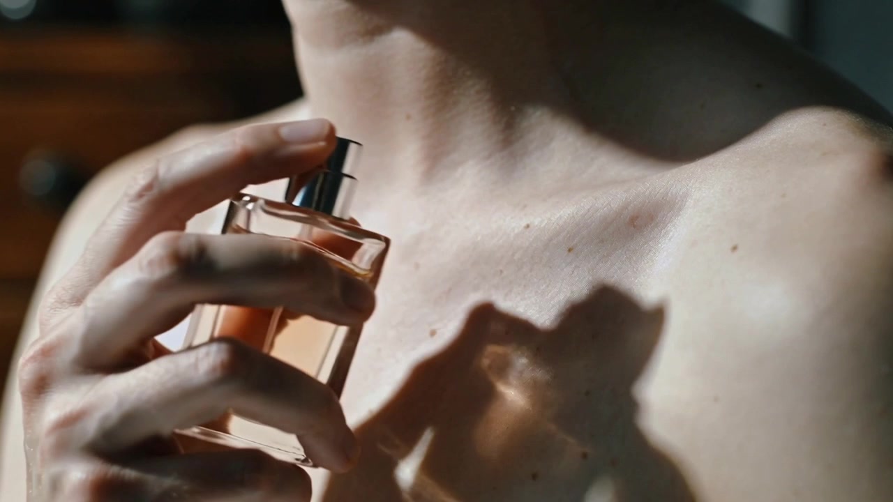 Donna con le spalle scoperte che solleva il profumo al collo vicino a una cassettiera, premendo l'erogatore per la fragranza.