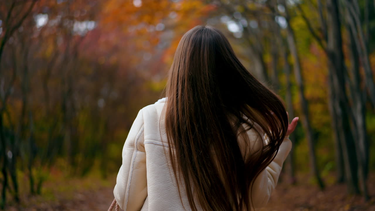 Long-haired brunette woman walking in the park. Lady turns around looks into camera holding a phone in her hand. Autumn nature backdrop.