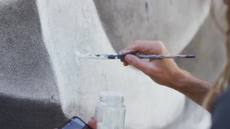 video de la sección media del artista masculino caucásico con dreadlocks usando pincel pintando mural en la pared