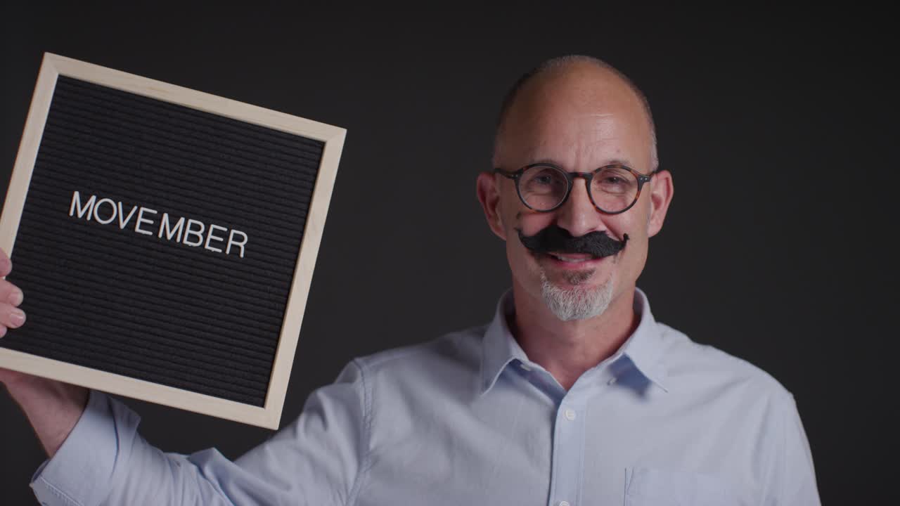 Portrait Of Mature Man Wearing Fake Moustache And Holding Sign Symbolizing Support For Movember Promoting Men's Health And Cancer Awareness