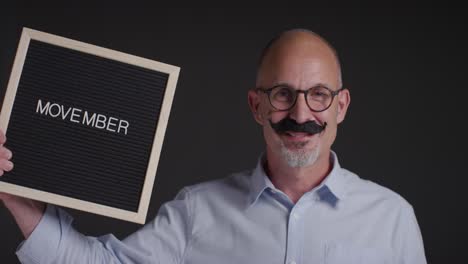 Portrait Of Mature Man Wearing Fake Moustache And Holding Sign Symbolizing Support For Movember Promoting Men&#039;s Health And Cancer Awareness