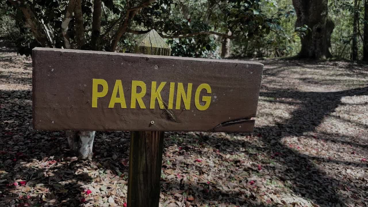 A wooden sign with yellow lettering reading “PARKING” stands along a shaded forest path surrounded by trees and fallen leaves