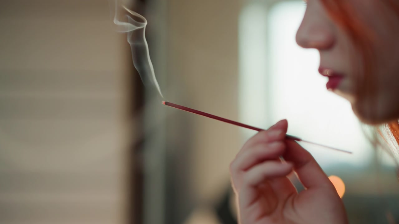 Nghi lễ thơm ngát thanh bình. Khói dịu nhẹ và ánh sáng ấm áp. Người phụ nữ dịu dàng thực hiện nghi thức thanh tẩy nhẹ nhàng bằng khói thơm. Cô ấy thở ra khói hương bên cạnh ánh sáng cửa sổ dịu nhẹ.