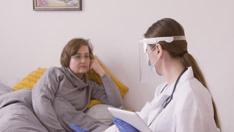 senior woman sitting on bed while talking to female doctor in medical mask and protective screen using a table