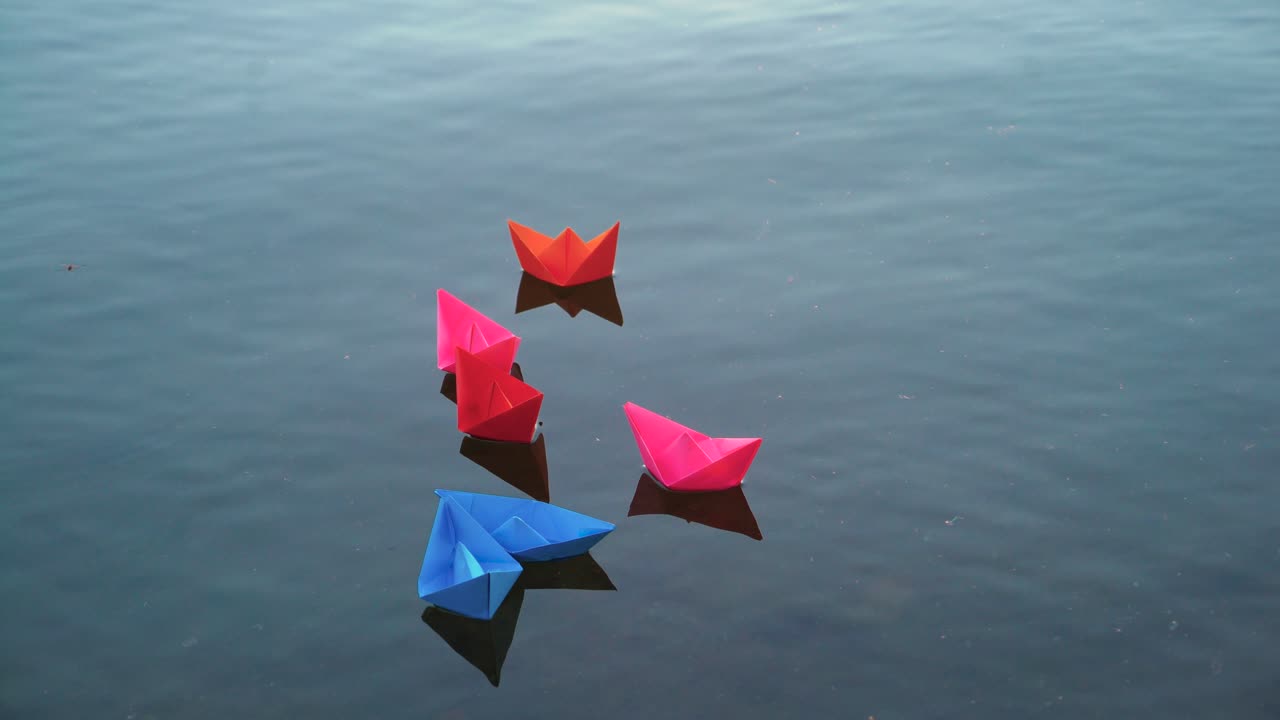 Six colored paper boats on blue water and some light splashes falling on. Beautiful children's origami ships floating in the river. Close-up