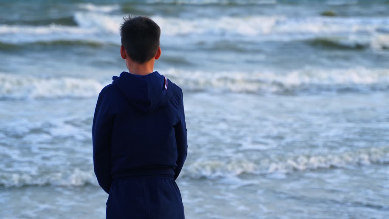 Back view of a boy near the blue sea. Teenage boy standing on the beach and looking on the sea water. Child dreaming on the ocean shore.