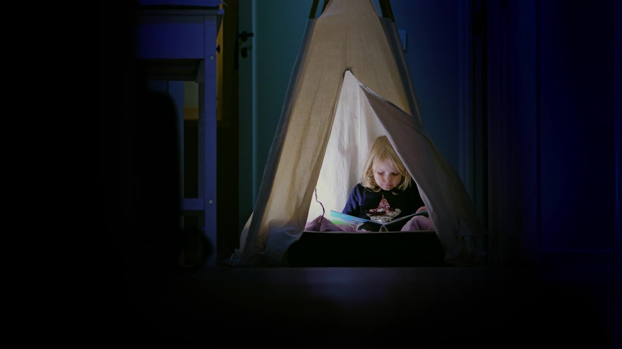 Cute little girl reading alone at night in a tent at home