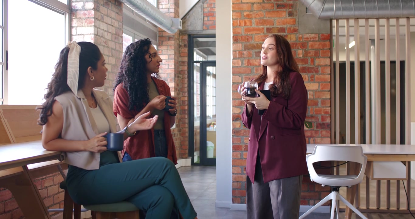 Three women in blazers at lounge, speaker gesturing with carafe making peers sipping mugs on break