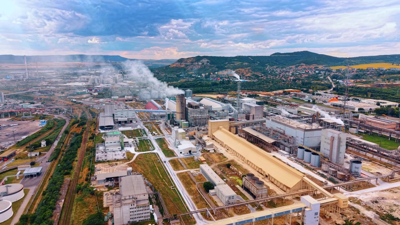 Industrial manufacturing plant with long warehouse buildings. Smokes rise from chimneys across a vast production area bounded by hills and a suburban settlement.