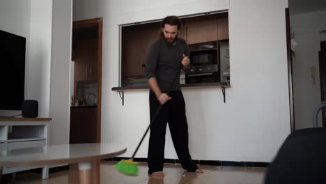 video de un hombre barbudo barriendo el piso de su sala de estar con una escoba negra y verde