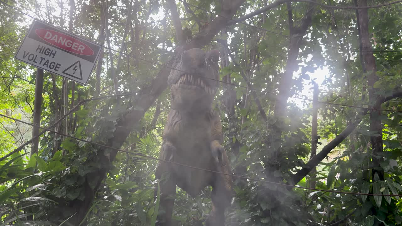 Animatronic dinosaur roars with mist effects near warning sign in lush, sunlit forest setting