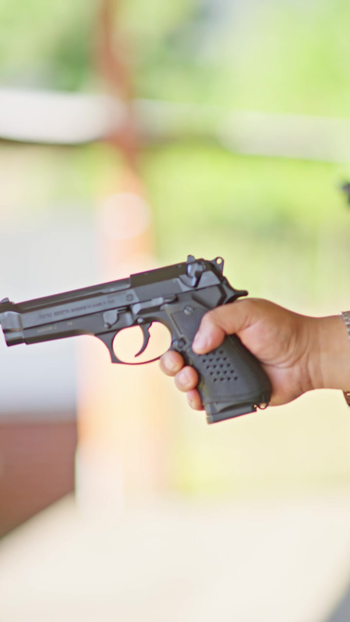 Close up of a hand holding a semi automatic pistol at an outdoor shooting range, with the covered firing line and green trees blurred in the warm background. Close up shot