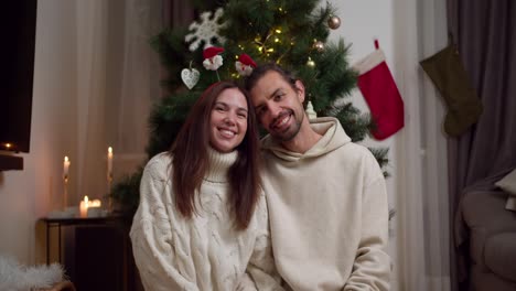 Portrait-of-a-happy-couple:-a-brunette-guy-in-a-white-sweater-and-a-brunette-girl-in-a-white-sweater-posing-and-looking-at-the-camera-near-their-New-Year-tree-in-a-cozy-winter-home