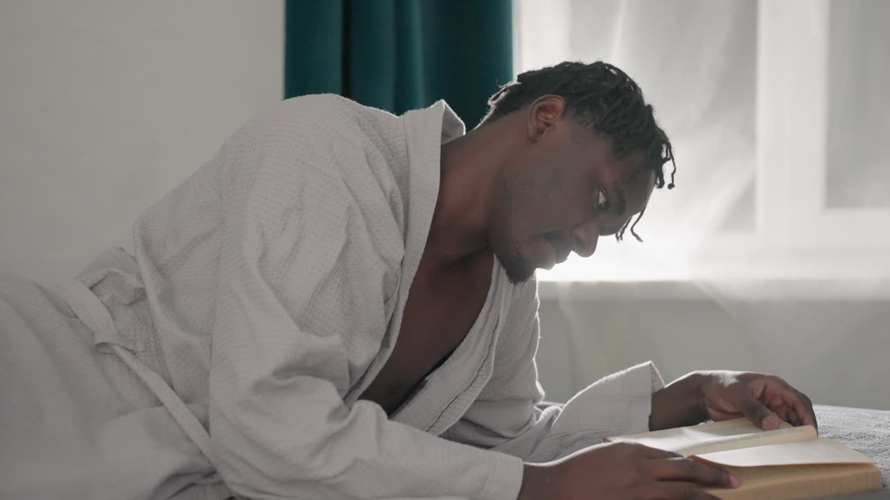 Champ in white bathrobe laying on bed, intently reading book with one hand, flipping pages, green velvet headboard in background, soft light from window creates a calm, peaceful atmosphere