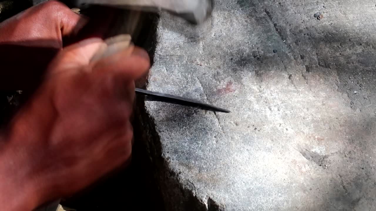 primer plano de la escena rural africana de una persona golpeando un trozo de metal con un martillo para convertirlo en una punta de flecha para cazar