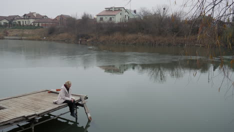 mujer sentada sola en un muelle junto a un lago congelado en un frío día de otoño o invierno