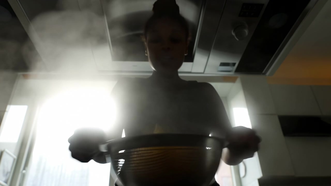A backlit person stands in a modern kitchen, holding a large metal bowl with rising steam as food preparation continues from an early to middle moment in the