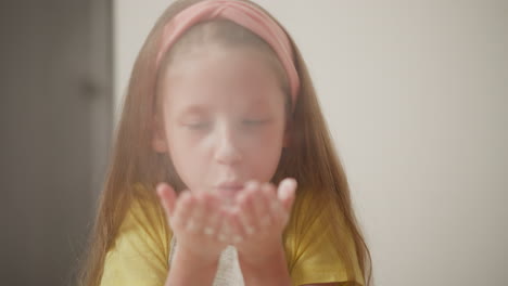 close up of girl in yellow shirt blowing flour from cupped hands as white powder creates soft cloud around her face in warm light, capturing playful and dreamy kitchen moment