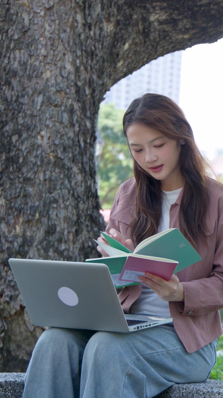 A student sits under a tree in a city park, reading and using a laptop