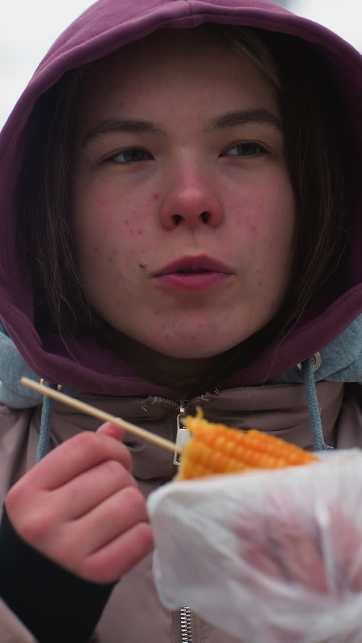 겨울 재을 입은 소녀가 거리에서 옥수수를 먹고, 거리 음식을 즐기고, 추운 날씨, 플라스틱으로 싸여, 도시 환경, 야외에서 캐주얼 식사, 옥수수, 후드 재, 음식 즐거움