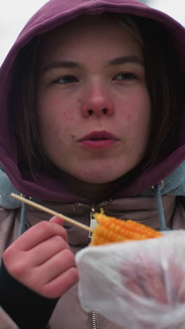 chica con chaqueta de invierno comiendo maíz en la calle, disfrutando de la comida callejera, clima frío, envuelta en plástico, entorno urbano, comida casual al aire libre, sosteniendo maíz, chaqueta con capucha, disfrute de la comida