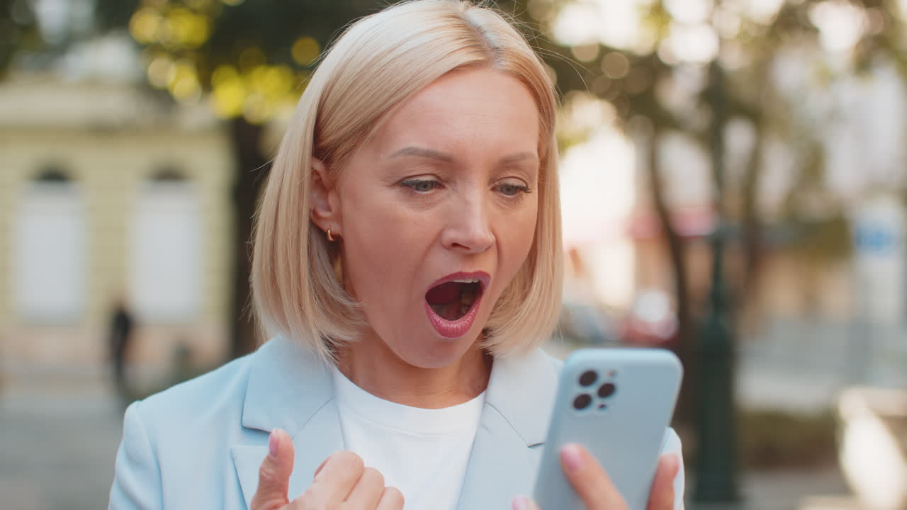 Una donna d'affari caucasica matura che sembra scioccata mentre legge buone notizie sullo smartphone in una strada della città