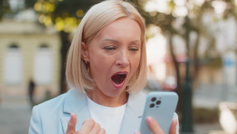 caucasian mature businesswoman looking shocked while reading good news on smartphone on city street