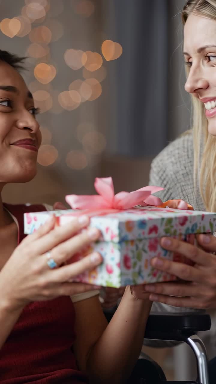 Video verticale: Donna bionda in maglione che si china per dare un regalo a un'amica in sedia a rotelle a casa, condividendo la gioia.