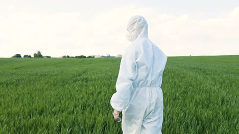 Vista-Trasera-Del-Investigador-Caucásico-Con-Traje-Protector-Blanco-Y-Gafas-Caminando-En-El-Campo-Verde-Mientras-Hace-El-Control-De-Plagas