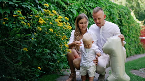 loving mom and dad sit squatted near their little son. cute blond baby boy drops the flower and picks it up.
