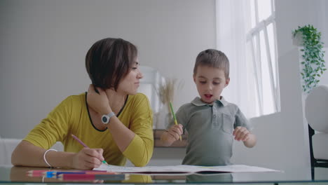 hermosa madre joven en un suéter cálido tendida en el suelo con mi hijo dibujando con marcadores en el papel retratando a su familia. el niño aprende a dibujar