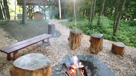 tilt fhd shot of a burning campsite fire surrounded by stumps for sitting and a private luxurious treehouse accommodation behind in dolní morava, czechia