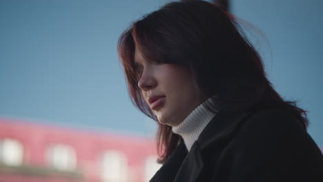 부드러운 갈색 머리카락과 사려 깊은 표정의 여성은 야외 환경에서 검은 코트와 색 터틀을 입고, 흐릿한 도시 배경과 은 파란 하늘을 배경으로