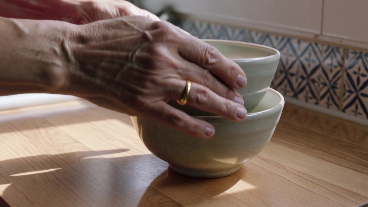 Mani di donna anziana che arrivano da sinistra e posizionano ciotole di ceramica sul bancone ad asciugare, con un anello visibile.