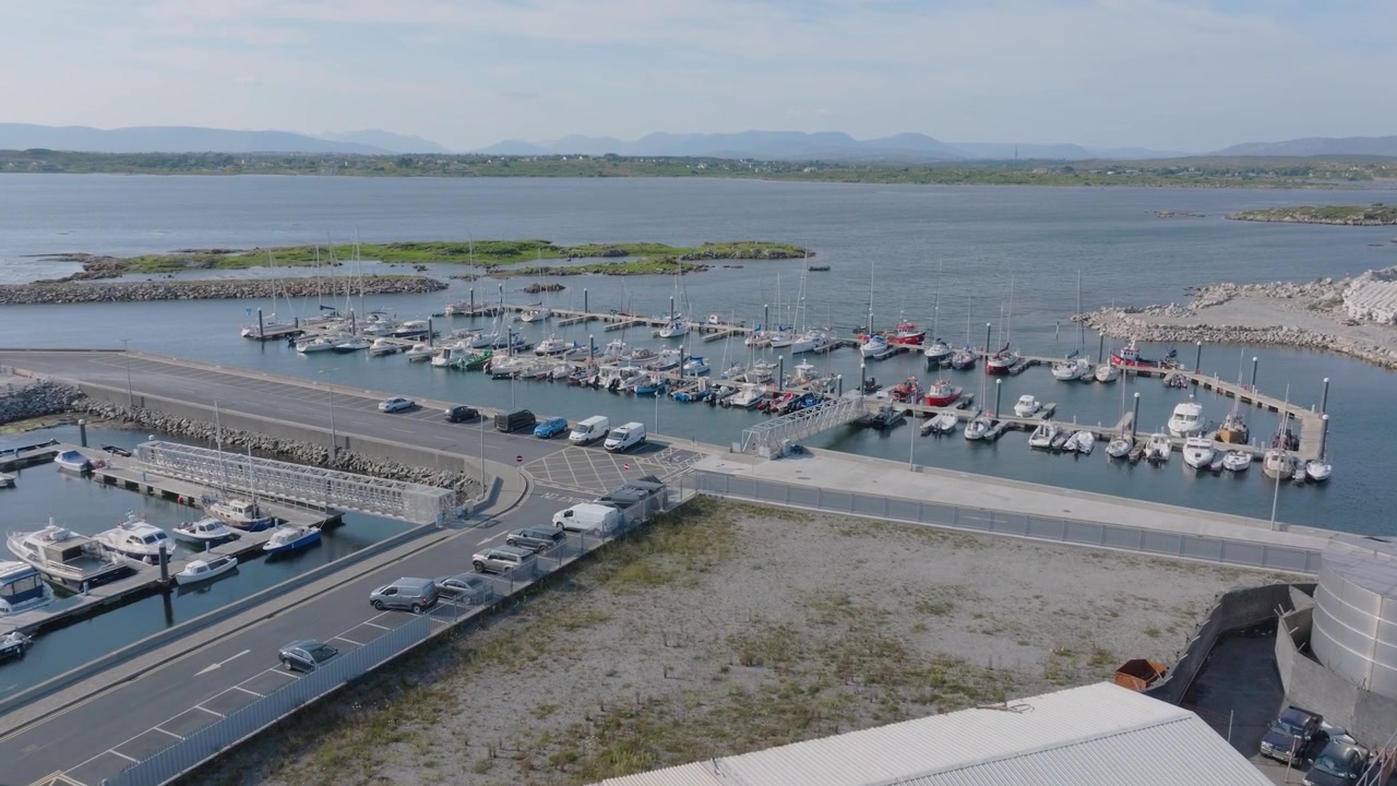 Yachts and motorboats fill Rossaveel boat berths beside breakwaters, access road and port buildings on a bright summer day.