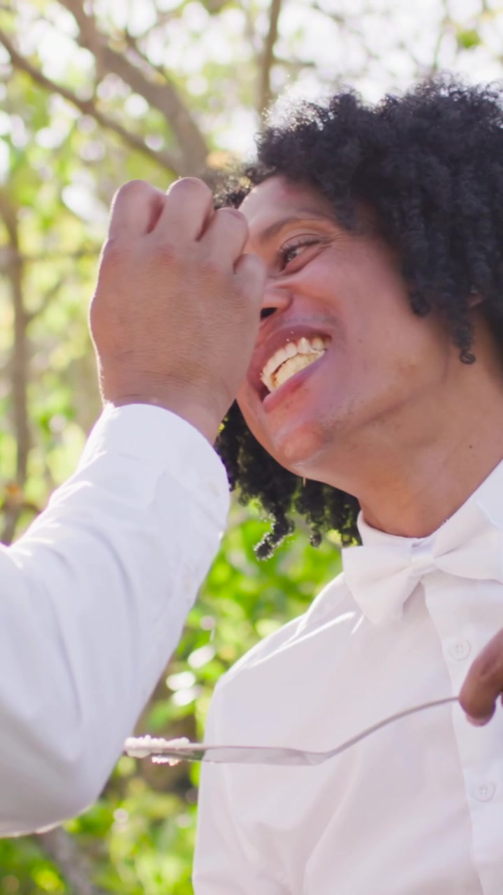 Video verticale: Uomo in camicia bianca che si protende per nutrire una donna che prende un morso di torta in giardino, condividendo.