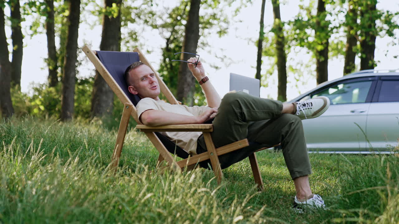 Man took a break from work and laid back in folding chair. Freelancer holds laptop and looks aside speaking, searching for ideas.