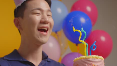 Close-Up-Studio-Portrait-Of-Man-Wearing-Party-Hat-Celebrating-Birthday-Blowing-Out-Candles-On-Cake