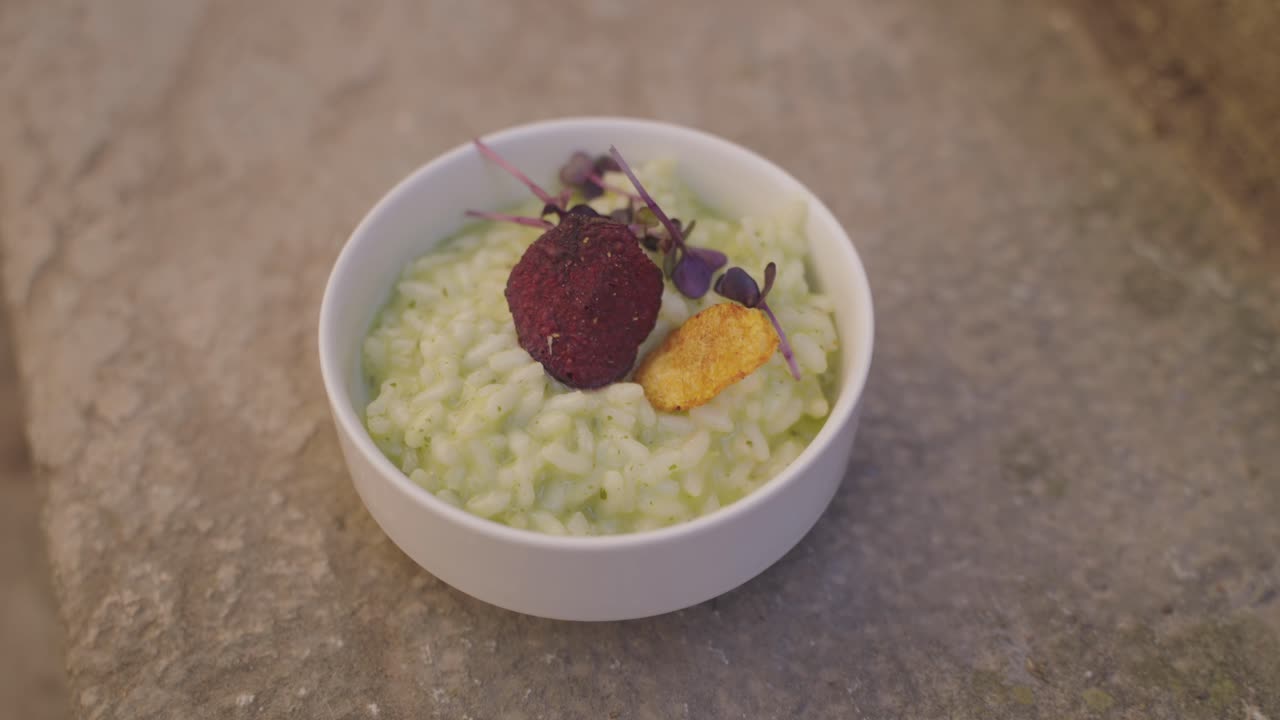 A beautifully plated bowl of creamy risotto garnished with colorful microgreens and crisp toppings on a rustic surface.